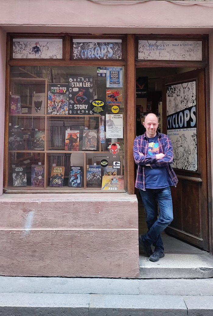 Photo d'un magasin de comic-book. On peut voir une grande vitre avec des titres en vente. Sur la droite, un homme se tient dans l'encadrement de la porte, c'est le vendeur du magasin. En haut de la vitrine il est marqué Cyclops, c'est le nom de la boutique.