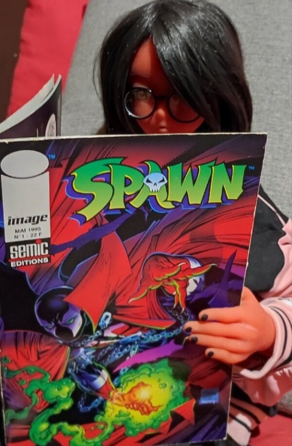 Image où Paprika, une poupée à la peau rouge, aux cheveux noirs et portant de petites lunettes rondes est assise, tenant le premier numéro du comic-book Spawn, ouvert entre ses mains.