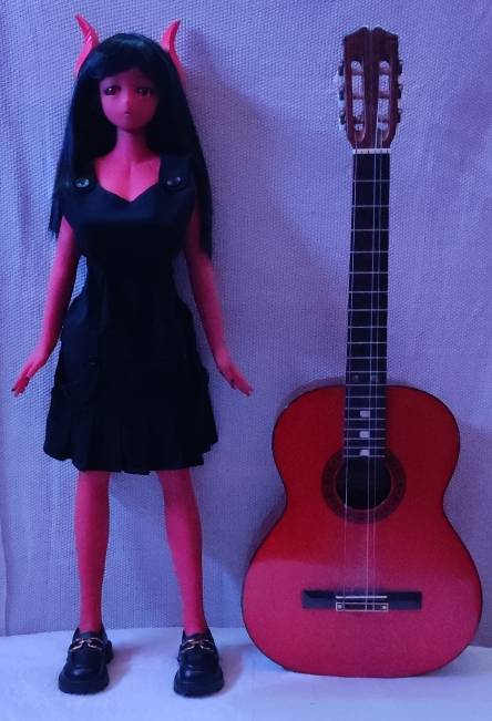 photo d'une poupée à la peau rouge, aux cheveux noires surmontés de deux petites cornes rouges. Elle porte une robe noire. Juste à coté d'elle il y a une guitare classique qui est posée, elles font toutes les deux la même hauteur.