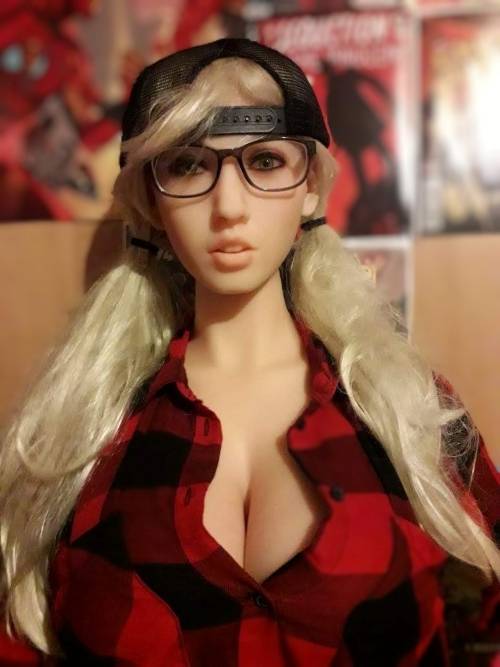 photo d'une love doll aux cheveux blonds. Elle porte une casquette à l'envers, des lunettes rectangulaires et une chemise à carreaux rouge et noire.