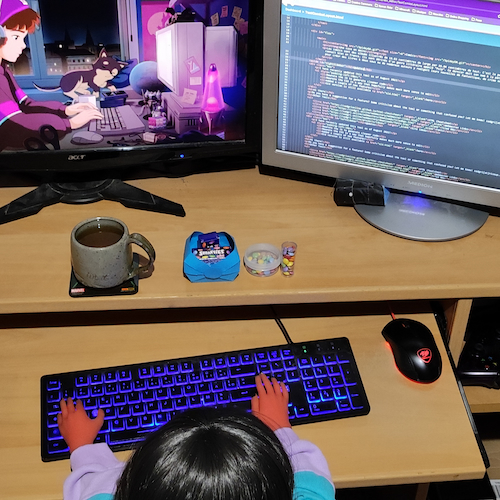 Photo d'une poupée aux cheveux noire assise à un bureau en bois clair sur lequel sont posés deux écrans d'ordinateur. Elle a également un clavier sous les mains ainsi que des smarties et une tasse de thé devant elle.