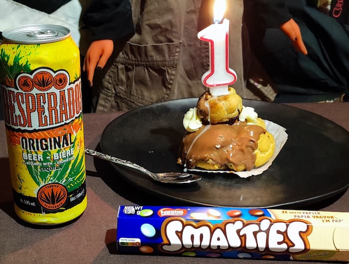 photo d'une assiette avec une religieuse au chocolat surmontée d'une bougie en forme de chiffre un. A côté de l'assiette il y a une canette de bière Desperados et un tibe de bonbons au chocolat smarties.