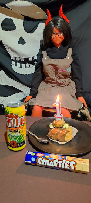 Photo d'une poupée à la peau rouge, aux cheveux noirs et aux yeux dorés. Elle est assise devant une table où il y a une assiette avec une religieuse au chocolat surmontée d'une bougie en forme de chiffre un. A côté de l'assiette il y a une canette de bière Desperados et un tibe de bonbons au chocolat smarties. Derrière elle un immense drapeau pirate fait office de fond.