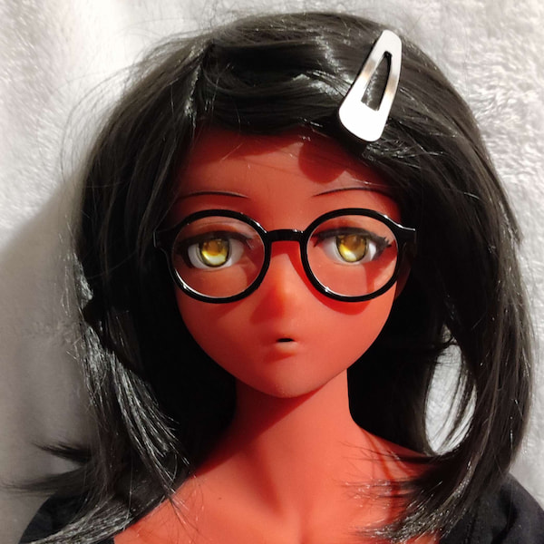 Photo en gros plan d'une poupée à la peau rouge, aux cheveux noirs et aux yeux dorés. Elle porte une petite paire de lunettes rondes à monture noire.